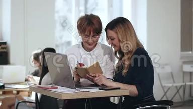 两位年轻漂亮的女经理与笔记本电脑和记事本一起工作，在<strong>现代</strong>阁楼<strong>办公桌</strong>上讨论工作。